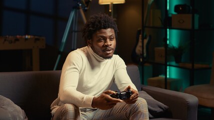 African american man sitting on couch at home, relaxing by playing videogames on gaming console. Gamer unwinding in apartment by competing in online multiplayer tournament
