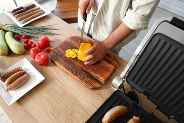 Young man cooking tasty sausages on modern electric grill and cutting fresh vegetables at table in kitchen