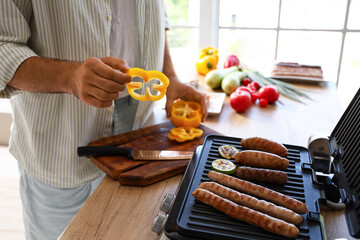 Young man cooking tasty sausages and vegetables on modern electric grill at table in kitchen