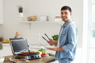 Young man cooking tasty sausages and vegetables on modern electric grill at table in kitchen