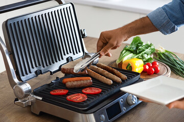 Young man cooking tasty sausages and vegetables on modern electric grill at table in kitchen