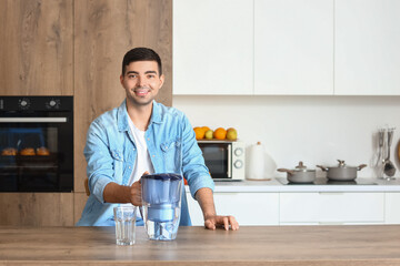 Young man with filter jug and glass of fresh water at table in kitchen