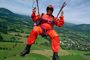 a man in signal red overalls hangs from a paraglider high above the meadows near Lenggries, pilot's...