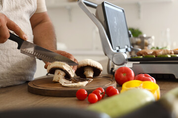 Mature man cooking tasty sausages on modern electric grill and cutting fresh vegetables at table in kitchen
