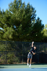 A young man plays tennis on a court surrounded by trees