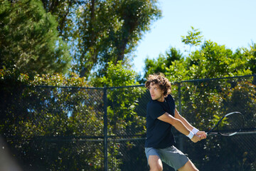 A young man plays tennis on a court surrounded by trees