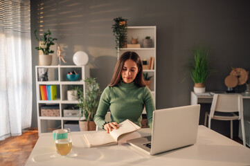 Smiling young woman taking notes while using laptop while working at home