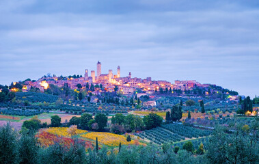 Obraz premium Town view of San Gimignano on a hill in the early morning at blue hour with cloudy sky in fall, all around are cypresses, olive groves and vineyards, Tuscany, Italy, Europe