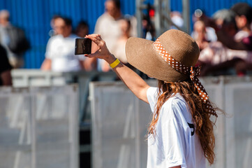 Woman in the sun wearing a wide-brimmed hat with an outstretched arm holding a cell phone and...