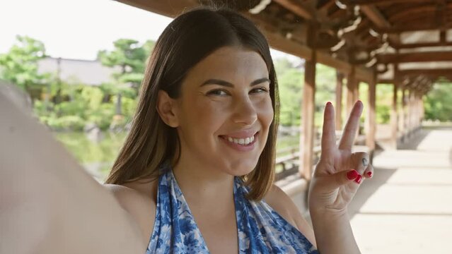Beautiful hispanic woman enjoying herself in kyoto, snapping a fun selfie at heian jingu, her dazzling smile and peace sign adding to her confident look