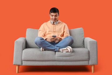 Handsome young man using mobile phone on grey sofa against orange background