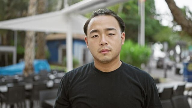 Defiant young chinese man, with a serious expression, stands tall on sunny terrace of urban coffee shop, resolutely shaking his head, saying 'no'. cool, casual lifestyle personified.