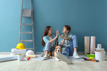 Young couple with paint brushes and Beagle dog giving each other high-five near blue wall during...