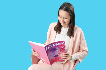 Young woman reading fashion magazine in chair on blue background