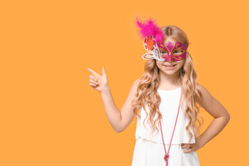 Funny little girl wearing carnival mask and pointing at something on yellow background