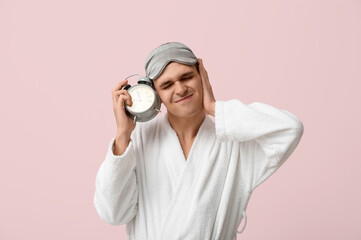Portrait of sleepy young man in bathrobe suffering from loud noise of alarm clock on pink background