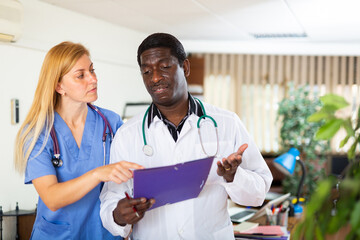 Two multinational doctors working at the clinic discuss work issues while standing in a well-lit...