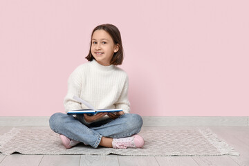 Little girl reading book while sitting on floor near pink wall