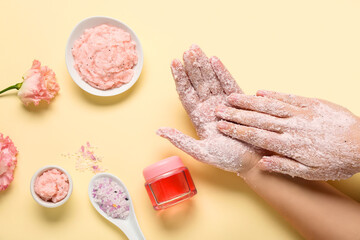 Obraz na płótnie Canvas Female hands with scrub, sea salt and cosmetic oil on light yellow background. Body massage concept