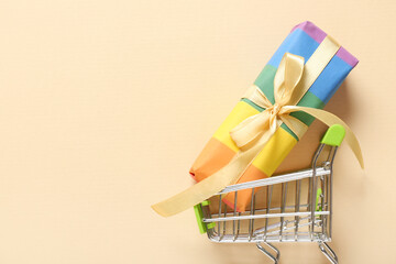 Small shopping cart with rainbow LGBT gift box on beige background. Valentine's Day celebration