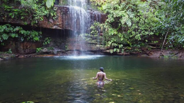 Woman in tiny bikini enters calm pool below pretty jungle waterfall