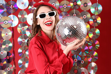 Beautiful young happy woman in stylish sunglasses with disco ball and compact disks garland on red...