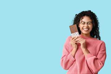 Happy young African-American woman with sweet chocolate bar on blue background