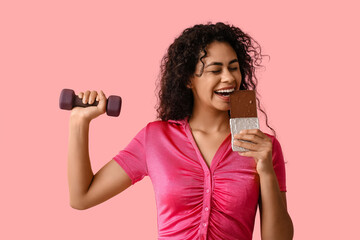 Beautiful young African-American woman with sweet chocolate bar and dumbbell on pink background