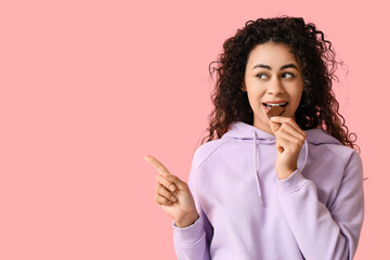 Beautiful young African-American woman eating piece of sweet chocolate and pointing at something on pink background