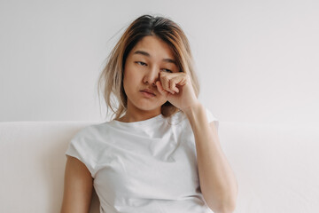 Sleepy face Asian woman yawning and rubbing her eyes in white t-shirt sits on the sofa.