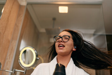 A joyful woman, with headphones in her ears, playfully dries her hair with a hairdryer in the bathroom after a refreshing bath, embodying a carefree and energetic moment of self-care and relaxation