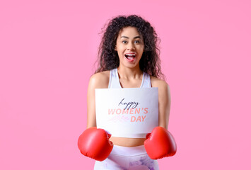 Portrait of young African-American woman in boxing gloves holding paper sheet with text HAPPY WOMEN'S DAY on pink background. International Women's Day celebration
