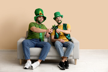 Young men in leprechaun hats with green beards holding bottles of beer and sitting on sofa near...