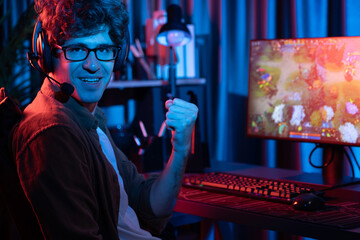 Host channel of young gaming streamer looking at camera to fist up in fighting Moba battle arena game with multiplay team against on blurred pc screen background at neon digital lighting room. Gusher. - obrazy, fototapety, plakaty