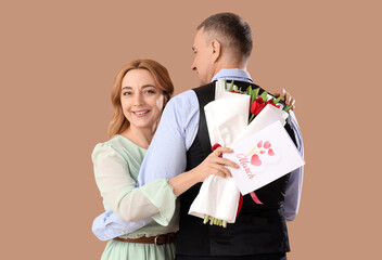 Middle-aged couple with bouquet of flowers and greeting card on beige background. International Women's Day
