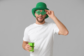 Young man in leprechaun hat and decorative glasses in shape of clover with glass of beer on grey...
