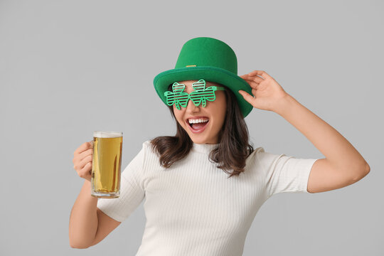 Beautiful young woman in leprechaun hat and decorative glasses in shape of clover with glass of beer on grey background. St. Patrick's Day celebration