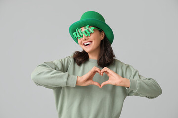 Beautiful young woman in leprechaun hat and decorative glasses in shape of clover showing heart...