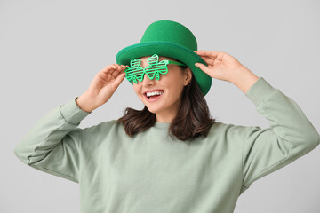 Beautiful young woman in leprechaun hat and decorative glasses in shape of clover on grey...