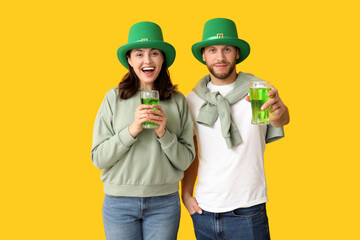 Happy young couple in leprechaun hats with glasses of beer on yellow background. St. Patrick's Day...