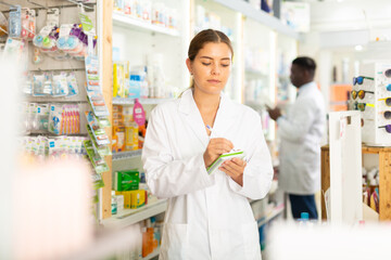 Portrait of a focused young female pharmacist standing in the sales hall of a pharmacy, making...