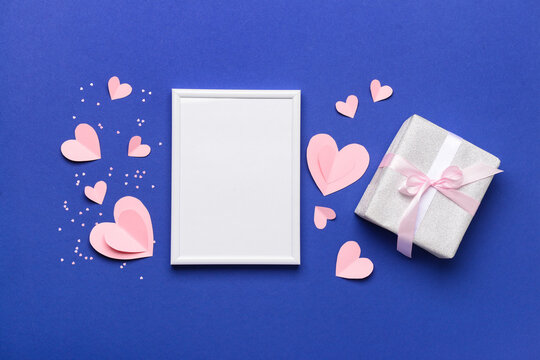 Composition with blank frame, gift box and paper hearts for Valentine's Day celebration on blue background