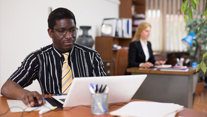 Focused african american man working for a large company types documentation on a computer while sitting at workplace ..in the office