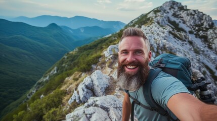 Naklejka na ściany i meble Hiker man reaches the summit of a mountain, capturing the triumphant moment by taking a selfie portrait. Convey the joy and accomplishment in his expression as he smiles at the camera