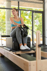 Fototapeta premium Girl stretching on reformer in gym. Caucasian young woman training pilates, using equipment.