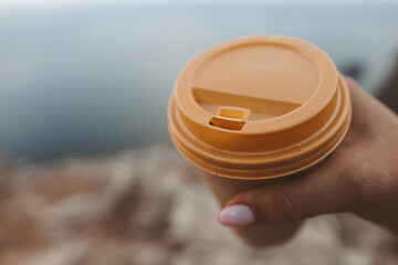 Hand holding Yellow cup with lid, coffee against a backdrop of a blue sky and sea. Illustrating cup...