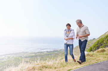 Holiday, walk and mature couple on road at the beach in countryside or nature with conversation of retirement. Summer, vacation and old man with woman talking on grass together at sea or ocean mockup