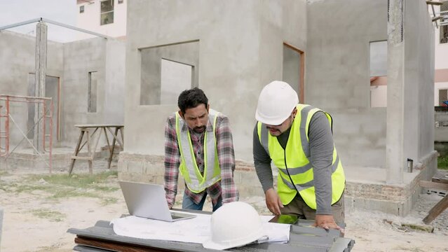 On-site workers discuss building plan. Asian engineers in white helmets and yellow vests work in team to build house with laptop and document. Unfinished cement house with ceiling joists construction