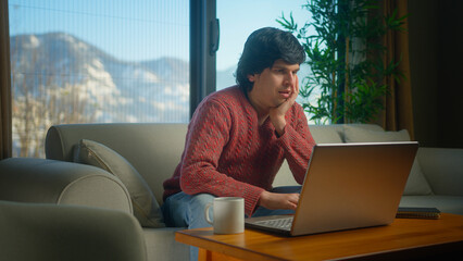 Distracted young college student working from home using with laptop while sitting on sofa in living room. Bored man student trying to study, staring blankly at the screen