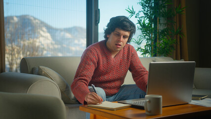 Young college student working from home using with laptop while sitting on sofa in living room at home. Focused millennial male looking at laptop, taking note on notebook